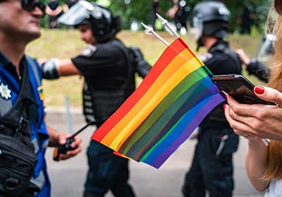 Person holding cell phone and rainbow flags near police